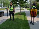 Children in the Montreal area became live speed cameras as a way to get drivers to slow down in school zones.  A policeman stands by as a child carrying a backpack equipped with a textbook showing the speed of drivers, Friday, June 30, 2023, handout photo.