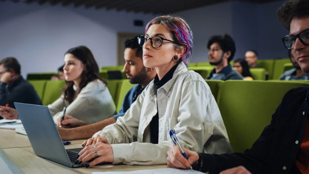 Photo of young adults with pink and purple hair, glasses, and white coats sitting in a classroom in front of a laptop